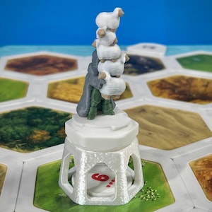 May include: A white and gray 3D-printed game piece featuring a figure holding a stack of sheep. The figure stands on a white hexagonal base with a die showing the number six. The game piece is set on a board with green, brown, and tan sections.