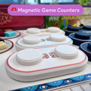 May include: Two white magnetic game counters with red and blue trim. The counters have a textured surface and are numbered 42. The counters are on a white surface.