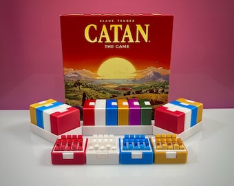 Può includere: La scatola del gioco da tavolo Catan presenta un'illustrazione di un paesaggio al tramonto. Sotto ci sono quattro set di pedine in plastica rossa, bianca, blu e gialla, organizzate in contenitori colorati e vassoi bianchi.