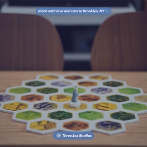 May include: A board game featuring a hexagonal grid of tiles in various colors including green, yellow, and brown. A small gray figurine stands on a central tile. The text "made with love and care in Brooklyn, NY" and "Three Sea Studios" are visible.