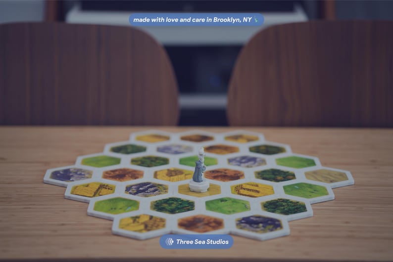 May include: A board game featuring a hexagonal grid of tiles in various colors including green, yellow, and brown. A small gray figurine stands on a central tile. The text "made with love and care in Brooklyn, NY" and "Three Sea Studios" are visible.