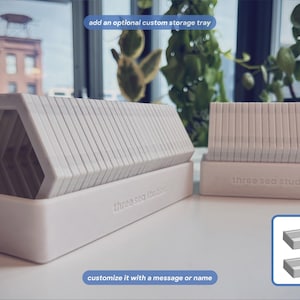 May include: Two white, rectangular storage trays with the text 'three sea studios' embossed on the front. The trays hold stacks of white, hexagonal-shaped tiles. A small inset shows two empty trays with the text 'customize it with a message or name'.