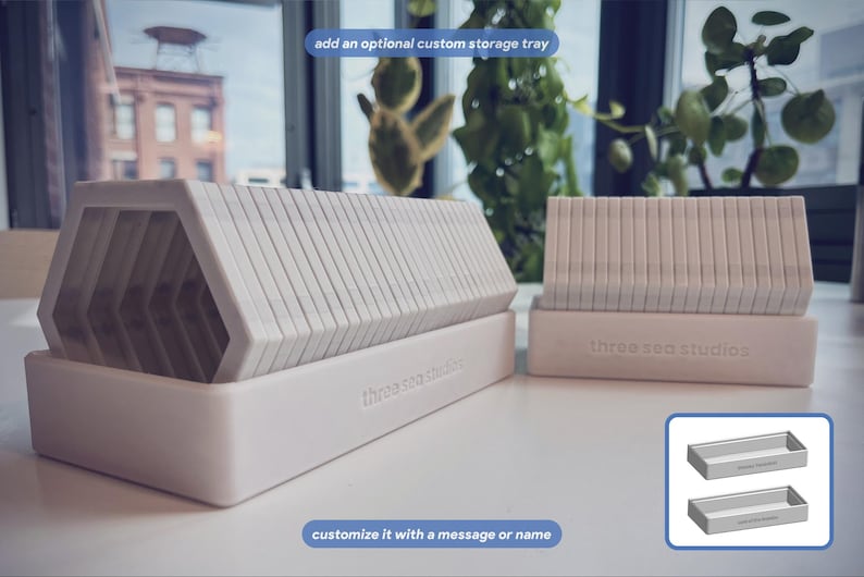 May include: Two white, rectangular storage trays with the text 'three sea studios' embossed on the front. The trays hold stacks of white, hexagonal-shaped tiles. A small inset shows two empty trays with the text 'customize it with a message or name'.