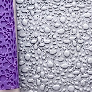 Könnte beinhalten: Eine violette zylindrische Walze und eine rechteckige graue Platte, beide mit einem sich wiederholenden geprägten Muster aus kleinen, abgerundeten Blättern an dünnen Stielen. Ein kleines blaues Etikett mit dem Text Digital files for 3D printing ist sichtbar.