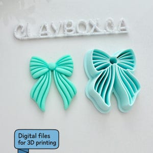 Op de afbeelding: Twee turquoise objecten in de vorm van een strik. E&eacute;n is een effen strik en de andere is een strikvormige uitsteker. Boven de strikken staat de tekst "CLAYBOX.CA". Daaronder staat een blauwe doos met de tekst "Digital files for 3D printing".
