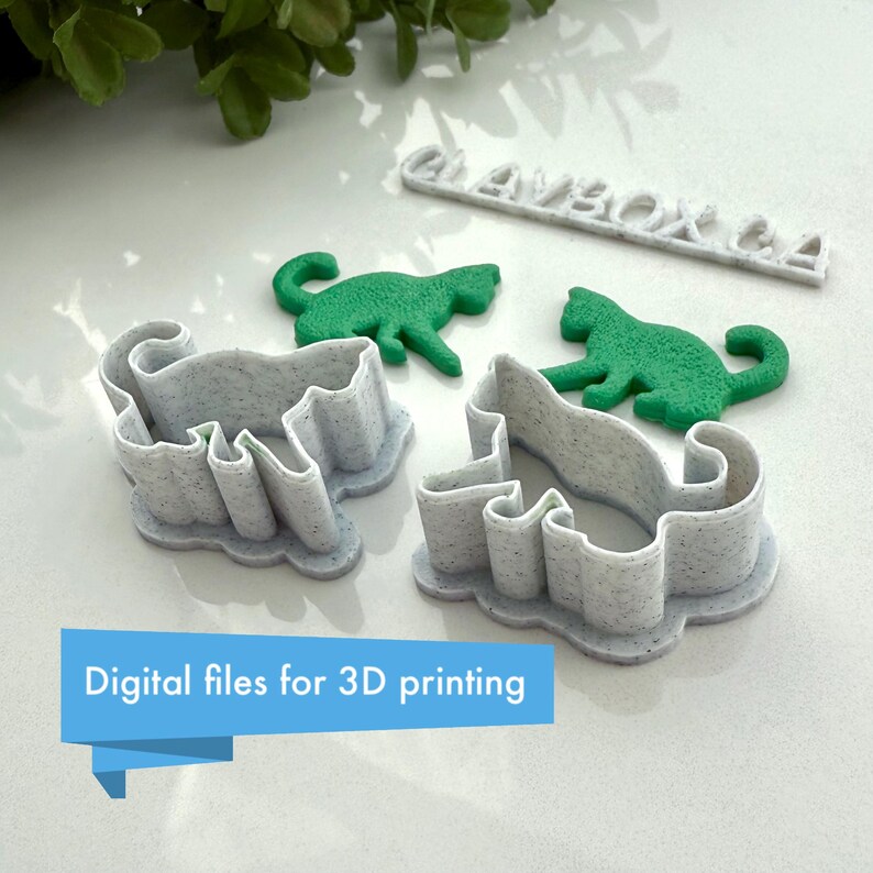 May include: Three 3D printed cookie cutters in the shape of cats. The cutters are white with a speckled texture. Two of the cutters are laying flat, while the third is standing upright. The cutters are on a white surface. The text "CLAYBOX CA" is printed on a white rectangular piece of plastic.