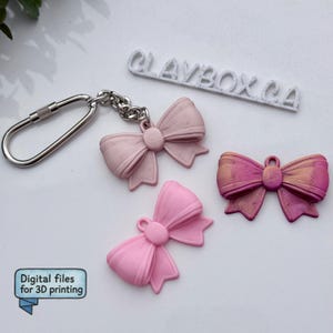 以下が含まれることがあります： ピンク色のリボン型のチャームが3つ。1つはキーホルダーに付いており、2つはバラバラです。1つのリボンは薄いピンク色、もう1つはゴールドのアクセントが付いた濃いピンク色、3つ目は無地のピンク色です。「CLAYBOX.CA」の文字が背景にあります。