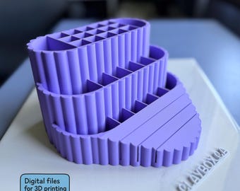 May include: A three-tiered, lavender-colored organizer with multiple compartments. The organizer has a fluted design on the sides and is labeled "Digital files for 3D printing". The base is a light color with the text "CLAYBOX CA".