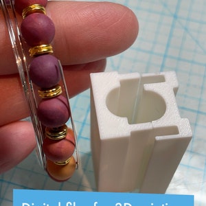 Op de afbeelding: Een witte 3D-geprinte mal voor het maken van armbanden. De mal is rechthoekig met een ronde opening in het midden. Naast de mal is een armband van rode, oranje en gouden kralen te zien.