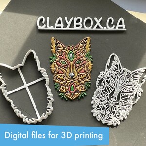 May include: Three 3D printed fox head cookie cutters. The centre fox is painted with gold, red, green, and blue paint. The other two fox cookie cutters are white. The text "CLAYBOX.CA" is printed in white on a black background.
