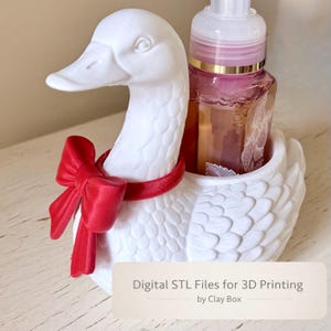 Könnte beinhalten: Ein weißer, entenförmiger Seifenspenderhalter mit roter Schleife und Kragen. Der Halter enthält eine transparente Flasche mit einem rosa-goldenen Etikett. Der Text "Digital STL Files for 3D Printing by Clay Box" ist sichtbar.