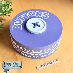 May include: A round, lavender-colored box with the word "BUTTONS" in white lettering on top, and a button-shaped lid. The box has a decorative white stitched pattern around the side. The text "Digital files for 3D printing" is in a blue box.
