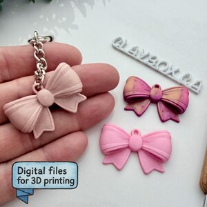 Può includere: Una collezione di modelli di fiocchi stampati in 3D in varie tonalità di rosa e un portachiavi. Un fiocco è attaccato a una catena d'argento. Il testo "Digital files for 3D printing" è in un riquadro blu. La parola "CLAYBOX CA" è visibile.