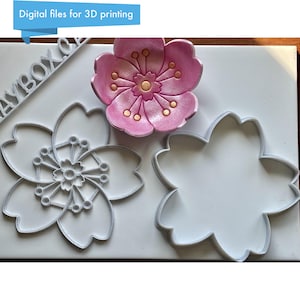 May include: A pink and gold cherry blossom, along with two white plastic flower-shaped cookie cutters. The image also includes the text "Digital files for 3D printing" in a blue banner.