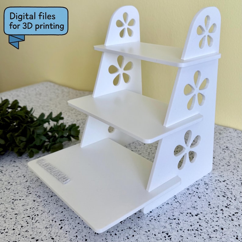 May include: A white, three-tiered display shelf with flower-shaped cutouts. The shelf has a rectangular base and two upright supports. The text "Digital files for 3D printing" is in a blue banner in the upper left corner.