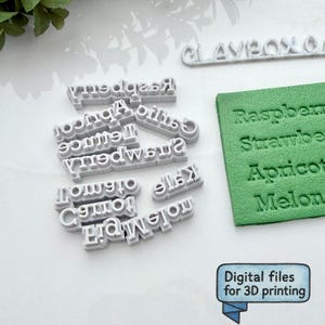 Puede incluir: Una colección de sellos de frutas y palabras grises impresos en 3D, incluyendo "Frambuesa", "Fresa" y "Albaricoque". Un bloque verde muestra texto estampado. La imagen también incluye el texto "Archivos digitales para impresión 3D".
