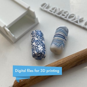 Puede incluir: Dos cuentas de arcilla polimérica azul y blanca, una con un patrón de remolino y la otra con un diseño floral. Las cuentas están sobre una superficie blanca con una herramienta de madera y una caja blanca etiquetada como "Claybox". Archivos digitales para impresión 3D.