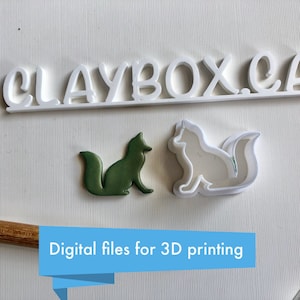 Puede incluir: Dos cortadores de galletas impresos en 3D, uno en blanco y otro en verde, con forma de zorros. Los cortadores están sobre una superficie blanca con el texto "CLAYBOX.CA" encima. El texto "Archivos digitales para impresión 3D" está debajo de los cortadores.