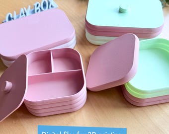 以下が含まれることがあります： ふた付きの3Dプリントボックスが2つ。1つのボックスはピンクで3つの仕切りがあり、もう1つは緑で1つの仕切りがあります。両方のボックスは丸みを帯びた正方形の形をしています。ボックスには白い文字で「CLAY BOX」とラベル付けされています。
