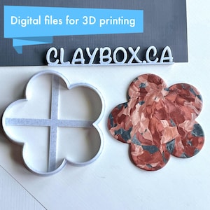 May include: A white plastic flower-shaped cookie cutter with a cross-shaped design inside. A red, black, and white marbled clay flower is next to the cutter. The text "CLAYBOX.CA" is visible above the cutters.