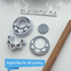 May include: A collection of gray polymer clay cutters and a wooden rolling pin. The cutters include heart shapes and a circular design. The text "CLAYBOX CA" is visible, along with the text "Digital files for 3D printing".