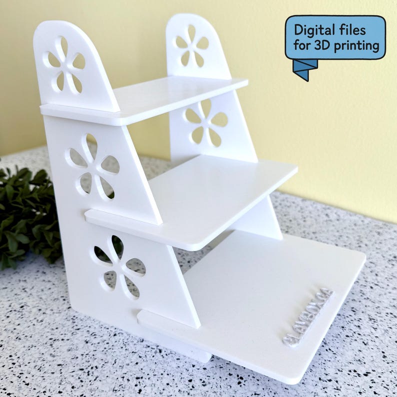 May include: A white, three-tiered display shelf with flower-shaped cutouts. The shelf has a stepped design and includes the text "Digital files for 3D printing" in a blue banner.