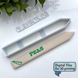 Op de afbeelding: Een lichtbeige tuinmarker met het woord "PEAS" in groen en een groene illustratie van een peul. Een lichtblauwe plastic mal ligt naast de marker. De tekst "CLAYBOX CA" staat boven de mal. De afbeelding bevat ook de tekst "Digitale bestanden voor 3D-printen".