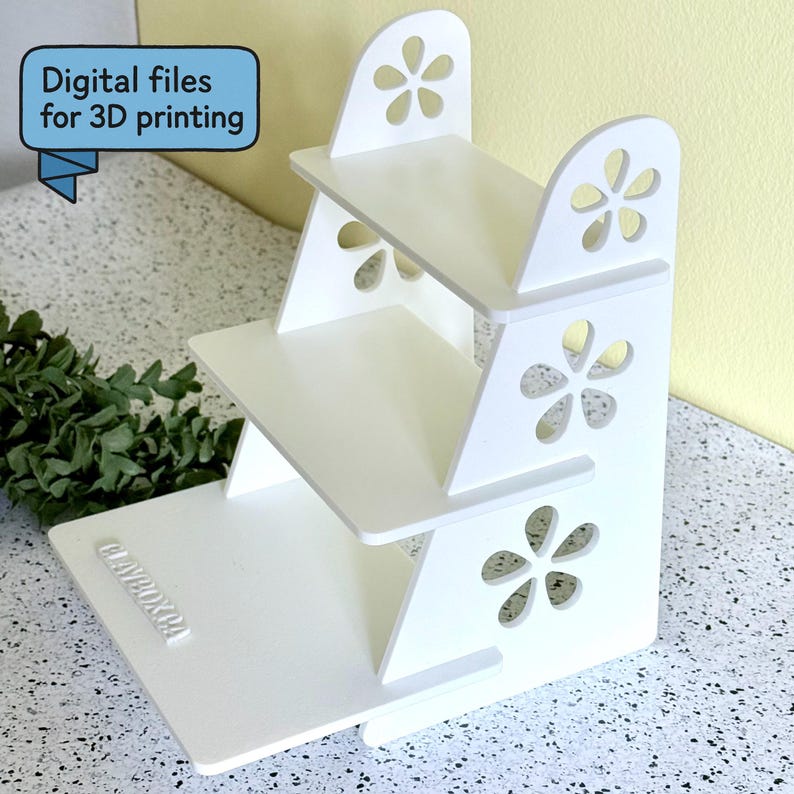 May include: A white, three-tiered display stand with flower-shaped cutouts and a rectangular base. The stand has a stepped design and the text "Digital files for 3D printing" is visible in a blue banner.