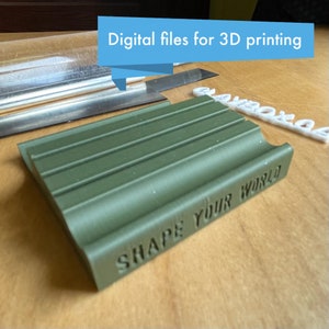 Puede incluir: Un jabonera verde impresa en 3D con las palabras "Shape Your World" en relieve en el lateral. La jabonera tiene cinco crestas en la parte superior para sujetar el jabón.