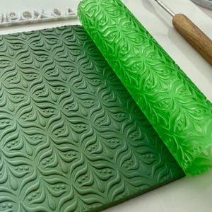 Puede incluir: Un rodillo verde con un patrón de hojas y puntos se muestra junto a una lámina de fondant verde con el mismo patrón en relieve.
