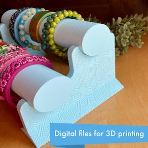 Peut inclure: Un présentoir à bijoux imprimé en 3D de couleur bleu clair avec deux tiges cylindriques pour tenir les bracelets. Le présentoir a un motif géométrique et est conçu pour exposer les bracelets.