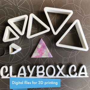 Puede incluir: Un conjunto de siete cortadores de galletas triangulares de plástico blanco de varios tamaños. Los cortadores están dispuestos sobre una superficie marrón con una pieza de arcilla triangular rosa y morada en el centro. El texto "CLAYBOX.CA" y "Archivos digitales para impresión 3D" es visible en la parte inferior de la imagen.
