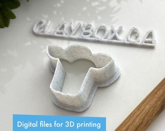 Op de afbeelding: Witte plastic koekjesvormer in de vorm van een koeienkop. De vormer heeft een gespikkelde textuur. De tekst "CLAYBOX.CA" is zichtbaar op de achtergrond.