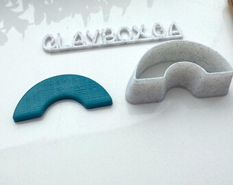 May include: Two 3D printed cookie cutters, one in white with a speckled finish and the other in teal blue. Both cutters are shaped like a half-circle arch. The white cutter has the text "CLAYBOX.CA" printed on it.