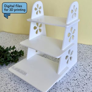 May include: A white, three-tiered display stand with a floral design cut into the sides. The stand has a rectangular base and the text "Digital files for 3D printing" in a blue speech bubble.