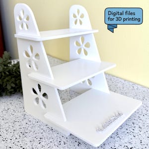 May include: A white, three-tiered display shelf with flower-shaped cutouts. The shelf has a stepped design and is made of a solid, opaque material. The text "Digital files for 3D printing" is in a blue banner. The shelf is suitable for displaying small items.
