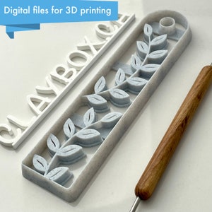 Puede incluir: Un letrero blanco impreso en 3D que dice "CLAYBOX.CA" con un molde gris impreso en 3D con un diseño de hojas. El molde está sobre una superficie blanca con una herramienta de madera.