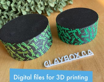 May include: Two black and green 3D printed round cake stands with a dragonfly design. The stands are on a wooden surface with the text "CLAYBOX.CA" and "Digital files for 3D printing" printed on the surface.