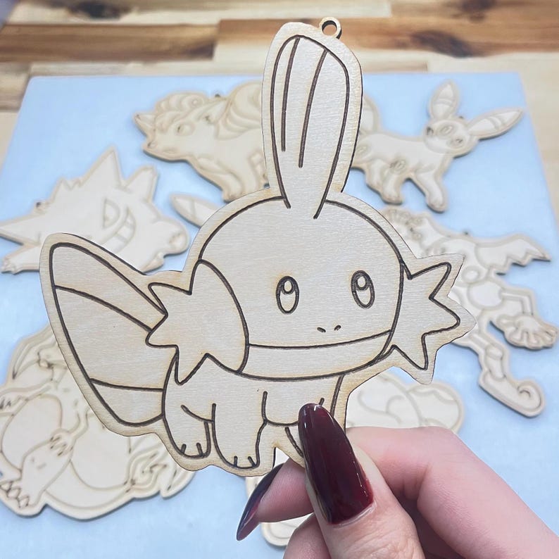 Close up photo of Mudkip wood cutout held in hand. Other characters are visible in the background, all made of light brown wood.