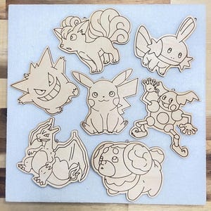 A collection of seven wooden cutouts featuring Vulpix, Gengar, Charizard, Pikachu, Fidough, Mr. Mime, and Mudkip. The cutouts are in a light brown color and have a smooth finish. Each cutout has a small hole at the top for hanging.