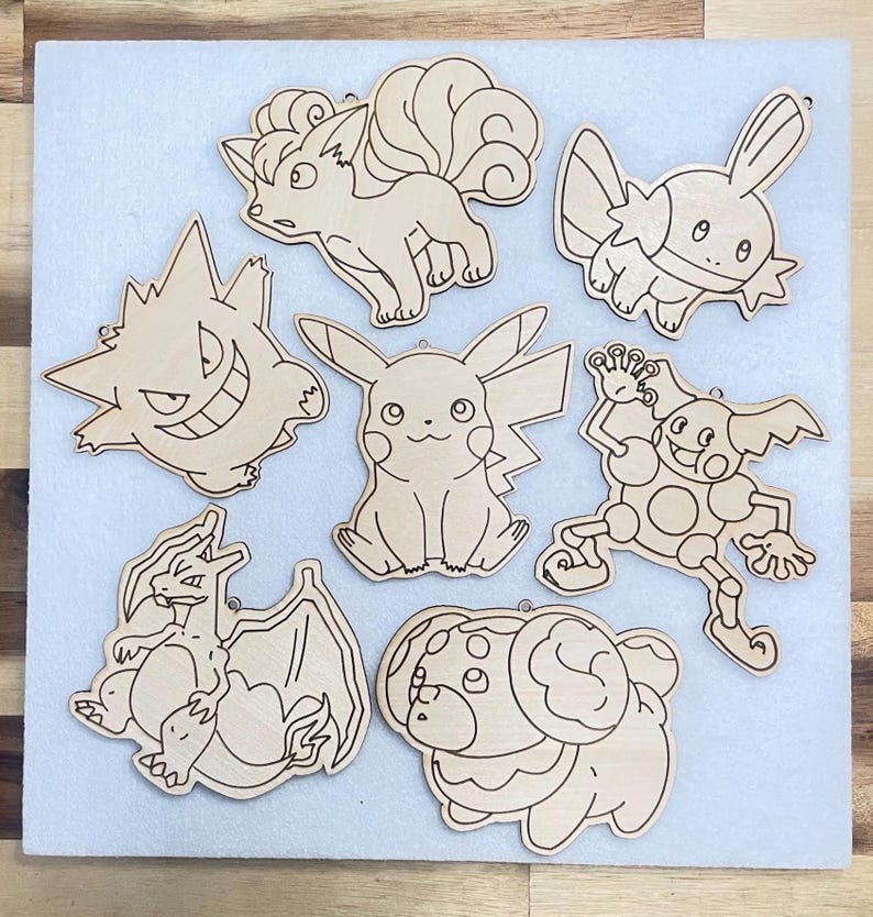 A collection of seven wooden cutouts featuring Vulpix, Gengar, Charizard, Pikachu, Fidough, Mr. Mime, and Mudkip. The cutouts are in a light brown color and have a smooth finish. Each cutout has a small hole at the top for hanging.