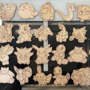 May include: A collection of wooden cutouts featuring various Pok&eacute;mon characters. The designs are laser-cut from light-colored wood, showcasing detailed outlines of each character. Includes a Pok&eacute;ball and several popular Pok&eacute;mon.