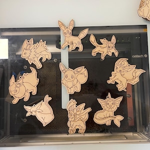 May include: A collection of wooden cutouts featuring various Pok&eacute;mon characters. Each cutout is a light brown color and has a small hole at the top for hanging. The designs include Eevee and its evolutions, showcasing detailed outlines of the characters.