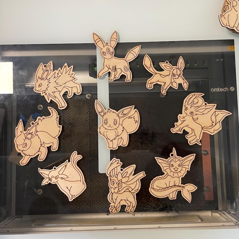 May include: A collection of wooden cutouts featuring various Pok&eacute;mon characters. Each cutout is a light brown color and has a small hole at the top for hanging. The designs include Eevee and its evolutions, showcasing detailed outlines of the characters.