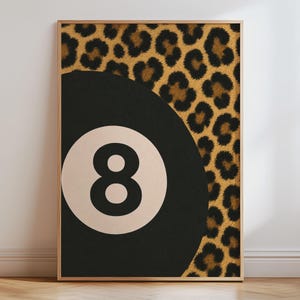 Puede incluir: Una lámina de arte mural rectangular en un marco de madera clara que presenta un diseño gráfico de una bola de billar negra con un centro circular blanco que contiene el número ocho, sobre un fondo con un patrón de estampado de leopardo.