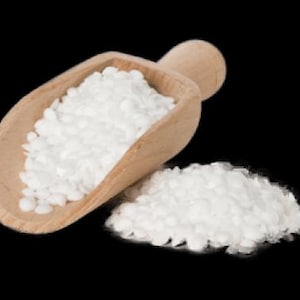 Può includere: Un cucchiaio di legno pieno di cristalli di bicarbonato di sodio bianchi, con un piccolo mucchio di bicarbonato di sodio che si riversa su una superficie nera.