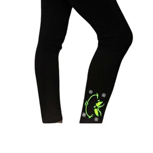May include: Black leggings with a neon green floral design on the lower leg. The design includes a curved stem with leaves and several small star-shaped accents. The leggings are made of a smooth, opaque fabric.