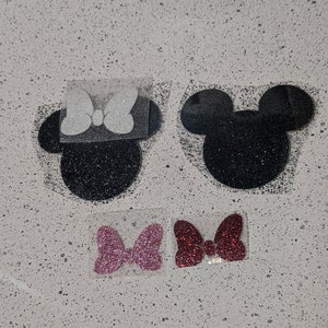 May include: Four iron-on patches featuring Mickey Mouse ears and a bow. Two patches are black with white bows, and two patches are pink with red bows.