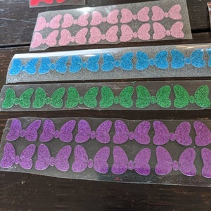 May include: Assortment of glitter bow tie stickers in various colors, including pink, blue, green, and purple. The stickers are arranged in rows on a clear sheet.