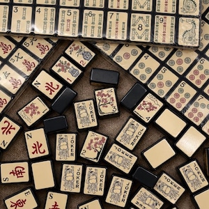 May include: A collection of cream-colored Mahjong tiles with black, red, and green ink designs, including numbered suits, directional characters, and Joker tiles. Some tiles are arranged in rectangular grids, while others are scattered loosely.